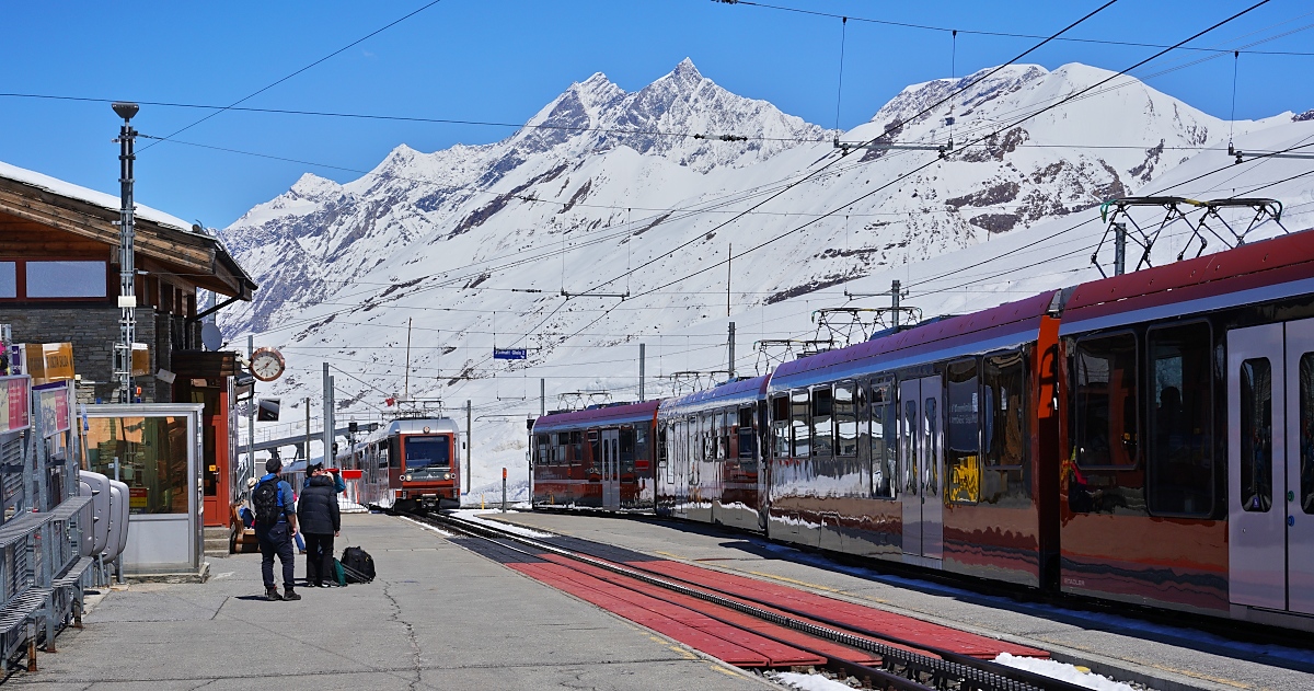Am 10.05.2024 kreuzen sich in der Station Riffelberg zwei Z�ge der Gornergratbahn: Im Hintergrund kommen der Bhe 4/6 3084 und ein weiterer Bhe 4/6 an, auf dem Nebengleis warten die Bhe 4/6 3094 und 3095 auf die Weiterfahrt nach Zermatt. In der Bildmitte die Mischabelgruppe mit dem Dom (mit 4545 m der h�chste Gipfel der Schweiz, der komplett auf schweizer Gebiet liegt) und T�schhorn (4491 m), rechts der Alphubel (4206 m). 
